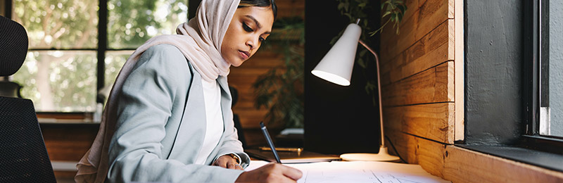 Female entrepreneur sitting at her desk writing out a promotional plan to create ads for her products and services.