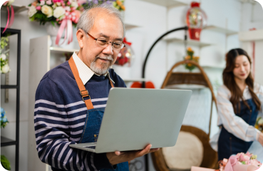 Older Asian-American entrepreneur sitting in his shop behind the front desk with his niece standing next to him. He holds a computer and is reviewing the complete outline of his business plan.